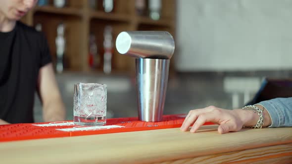Cocktail Shaker and Glass with Ice Standing on Bar Counter with Blurred Bartender Working at