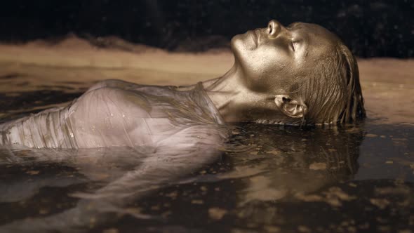 a Woman with Dyed Gold Skin and Hair Lies in the Water Posing