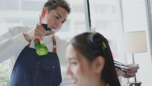 Asian professional male hairstylist combing and using scissors cutting woman's hair in salon.