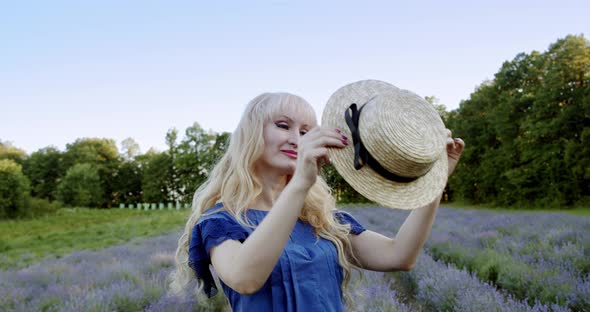 A Woman Stands on a Lavender Field and Puts a Straw Hat on Her Head on Summer Day. Slow Motion