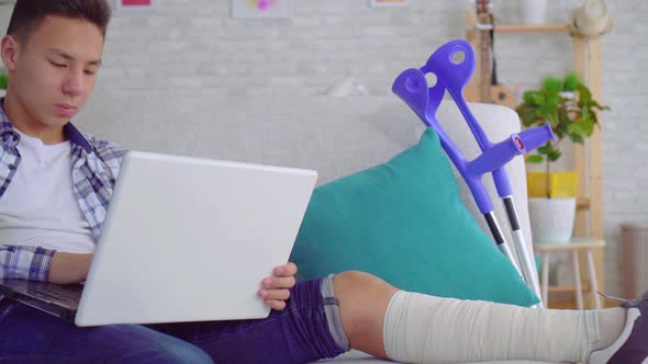 Asian Young Man with Broken Leg in Bandages Lying on Sofa with Laptop Close Up