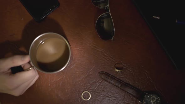 Mans Hand Holding A Cup Of Brown Coffee, Watch, Sunglasses, And A Pair Of Rings On The Brown Table -