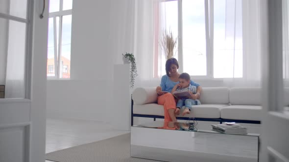 Loving Mother Teaching Her Son To Read Holding Book Speaking On Couch At Home