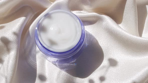 Blue glass cream jar on smooth silk background in sunlight with flowers shadow 4K skincare cosmetics