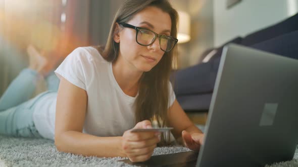 Woman with Glasses Is Lying on the Floor and Makes an Online Purchase Using a Credit Card and Laptop