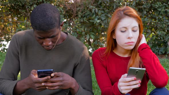 Young mixed couple sitting on grass using smartphones on their own