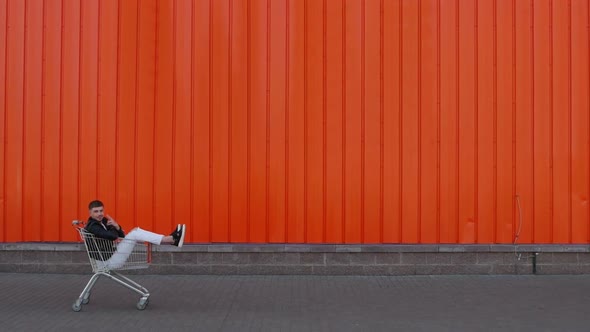 Happy Customer Man Is Sitting in a Shopping Cart and Rides Along the Wall of Mall Store Center