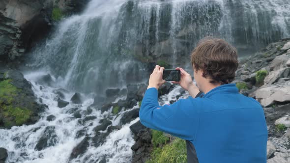 Man Holds Phone and Makes Video Standing Near Waterfall