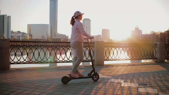 Attractive Woman Dressed in Pink Jacket Rides an Electric Scooter on Promenade