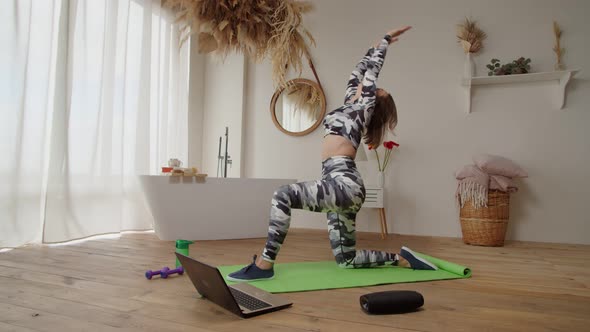Pretty Sporty Fitness Female with Laptop on Exercise Mat Doing Crescent Moon Pose Indoors