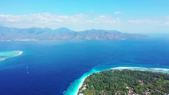 Aerial of bright blue waters between a beautiful beach island and distant land with scenic mountains