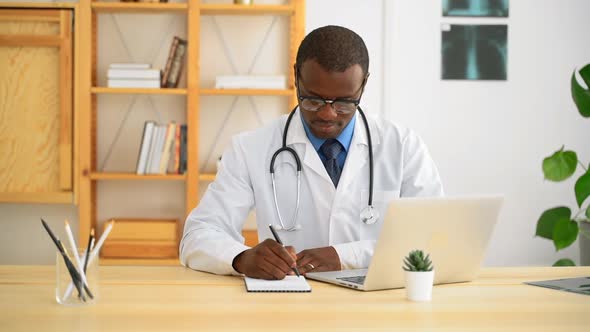 Young Male African Doctor Writing in Notebook and Working with Laptop at Table in Clinic Spbas