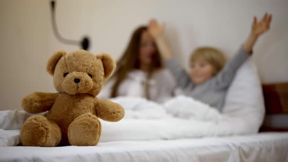 Closeup of Teddy Bear with Blurred Mother and Son Lying in Bed Talking and Gesturing