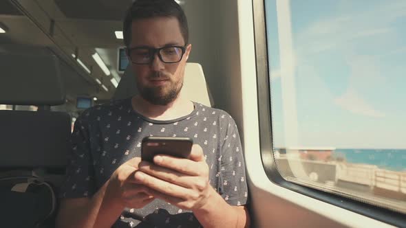 Bearded Young Man Is Sitting in Wagon of Suburban Train, Using Smartphone