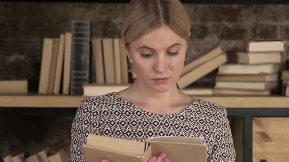 Close up of the Caucasian young beautiful woman student in glasses reading a book among books 