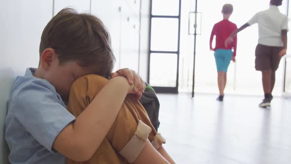 Video of sad caucasian boy sitting at school corridor, covering head with hands