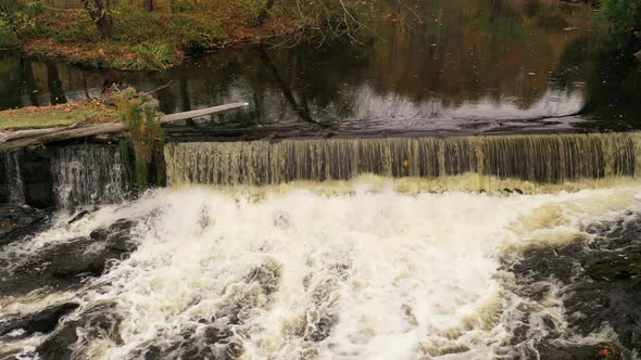 An drone view of a still lake which flows to a waterfall surrounded by colorful fall foliage in upst