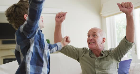 Happy caucasian grandfather and grandson jumping and raising hands in living room