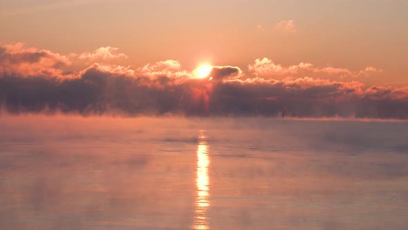 Timelapse of the sun rising through clouds over a very cold lake with steam rising in the winter tim