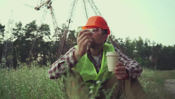 Hungry Worker in Overalls in Electric Power Business Eats Sandwich and Drinks Takeaway Coffee at