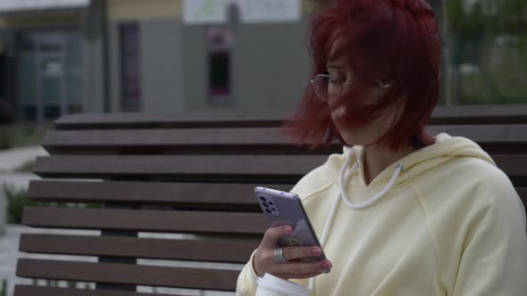 Young redhead Ukrainian teen girl with mobile phone sit on bench on city street