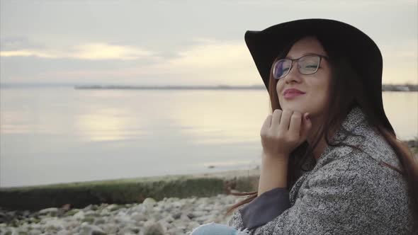 Young Pretty Smiling Woman in Black Hat and Glasses Sitting Near Sea at Sunset