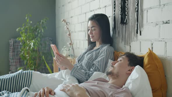 Asian Woman Using Smartphone Touching Screen Smiling While Biracial Race Man Sleeping in Bed at Home