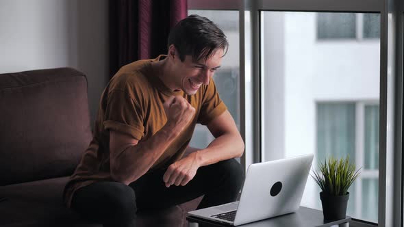 Young Man Sitting on Sofa at Home and Using Laptop Gets Surprised Then Happy and Celebrates Win