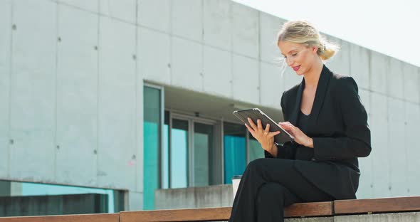 Side viewYoung Smiling Attractive Woman in Dark Tail Coat is Sitting Outside and Uses Tablet