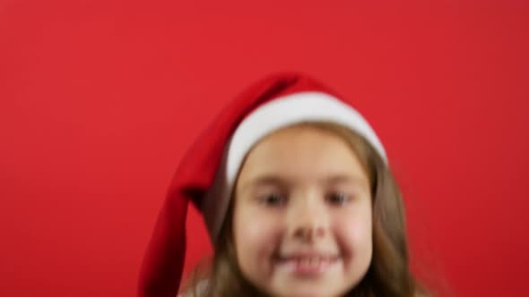 Smiling Girl in Santa Hat Pointing to the Side. Red Background With Blank Space
