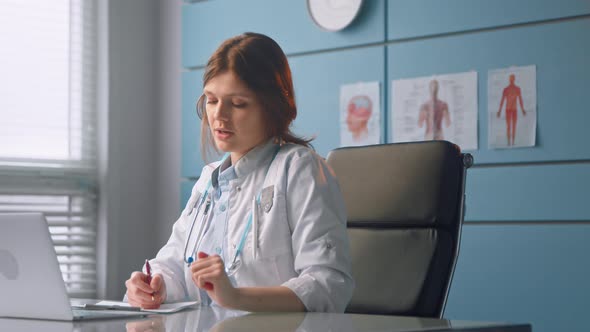 Young woman doctor talks to patient looking at sheets of papers and grey laptop display