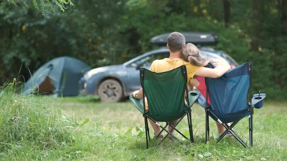 Happy couple sitting on chairs at campsite hugging, talking happily together and a car