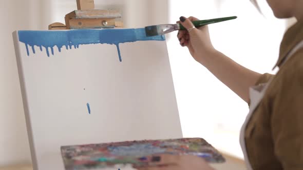 Woman is painting with brush on a canvas. Splitting the blue paint over the top edge of canvas.