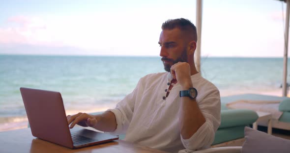 Bearded Young Man Working at Computer While Sitting at Table on the Open Verandah of the Beach Cafe