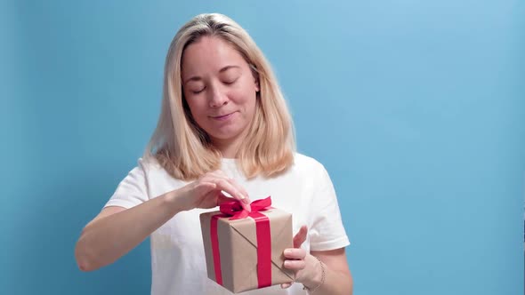 Young Smiling Woman Holding Gift Box and Gives It By Hands to Camera on Blue