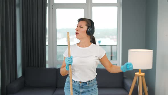 Woman in Headphones Cleans the House and Have Fun Singing with a Broom Like a Star at a Concert