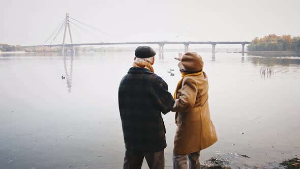 Loving Aged Spouses Talking Standing on Bank of River and Feeding Ducks Floating in It Park on