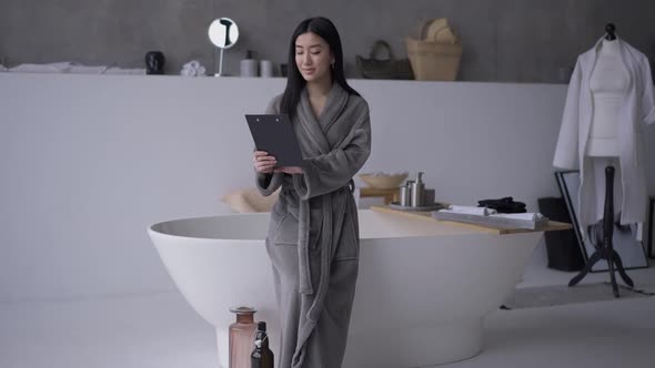 Wide Shot Portrait of Slim Asian Woman in Bathrobe Standing in Bathroom with Documents