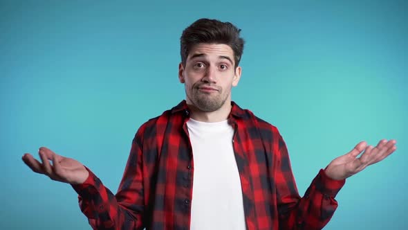 Young handsome unsure guy in red plaid shirt shrugs his arms, makes gesture of I don't know