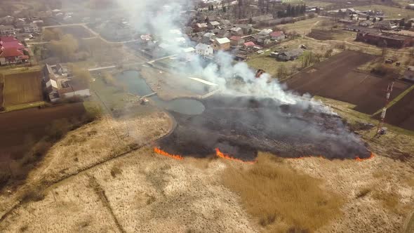 Aerial view of a field with dry grass set on fire with orange flames and high column of smoke.