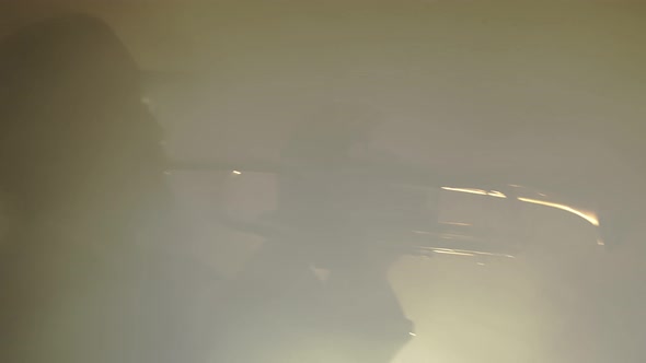 Bearded male musician in hat and jacket plays trumpet, pressing keys, closeup in smoke studio