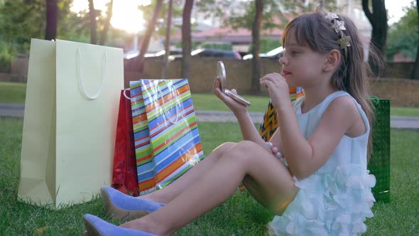 Glamorous Kid with Red Lipstick Doing Makeup and Looks Into Mirror in Hands Outdoors After Visit of