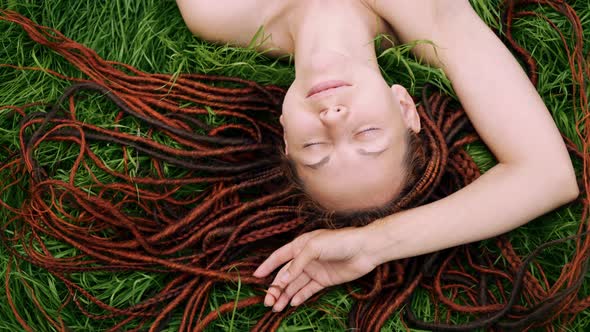 girl with red dreadlocks lying on the green grass, squinting from the sun, smiling. view from above