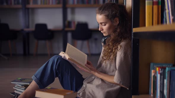 Student in Headphones Sitting Against Bookshelf with a Books on the Floor Closeup