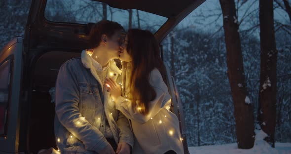 Young Couple Sit in a Car Trunk Surrounded By Christmas Lights Embracing