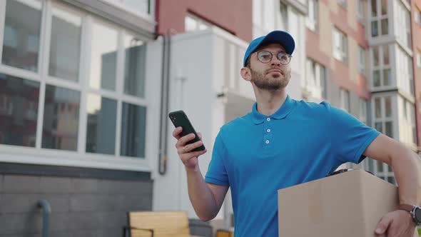 Caucasian Young Pretty Man Delivery Worker in Blue Cap Walking the Street and Carrying Carton Box