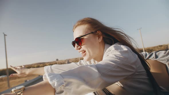 Happy woman in sunglasses rides convertible leaning out window with her arms up