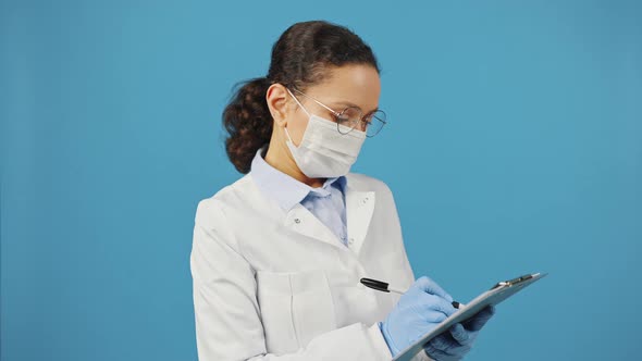 Medical Doctor in Protective Mask and Eyeglasses Writing in Clipboard and Looking at Camera Blue