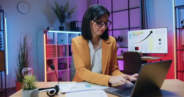 Brunette in Stylish Wear and eyeglasses sitting in front of computer and working