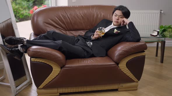 Desperate Frustrated Young Asian Groom Lying on Armchair Holding Glass of Whiskey